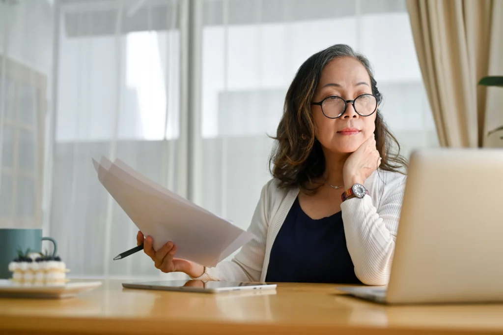 Professional woman sitting at desk with documents in hand while looking at laptop. She has a rainbow coloured watch to left wrist