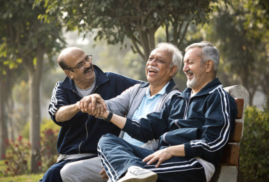 Three elderly men sitting on bench laughing and holding hands with trees to reverse