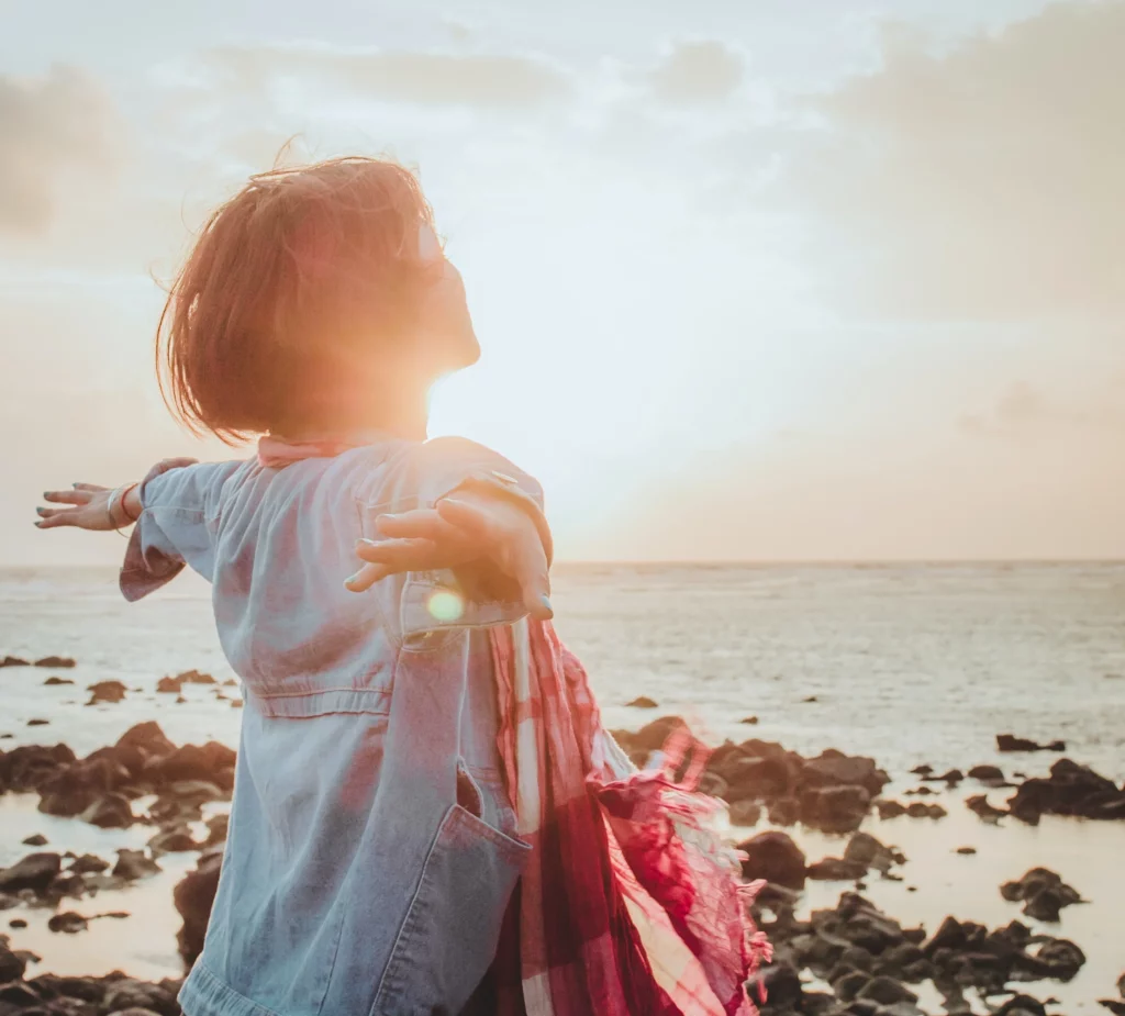 Woman standing by shoreline with arms open and sunlight to face