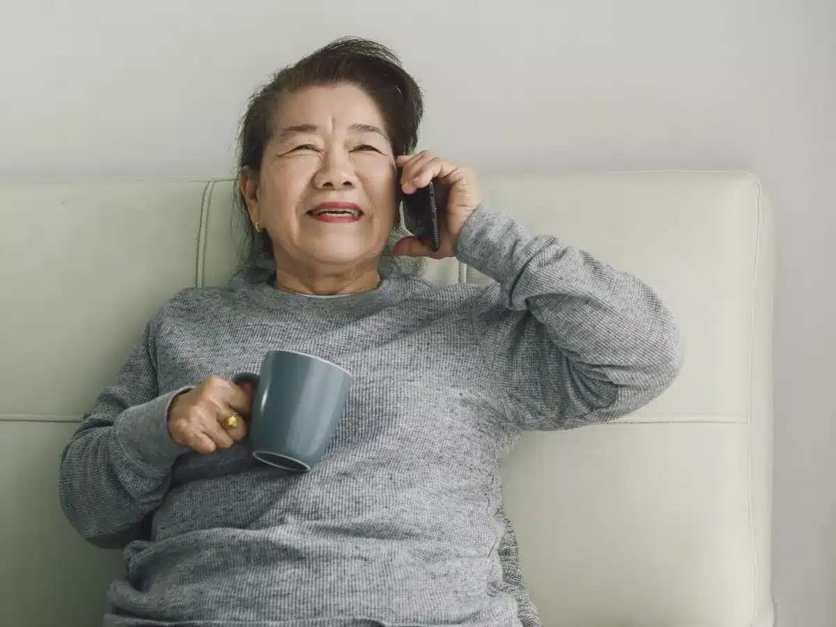 Asian woman smiling and holding a mobile phone to her ear while also holding a mug to her chest