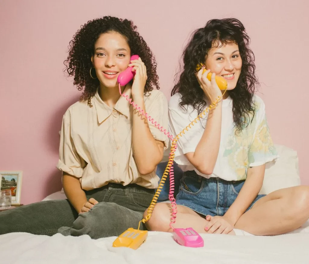 2 female friends sat next to each other holding vintage telephones to their ears