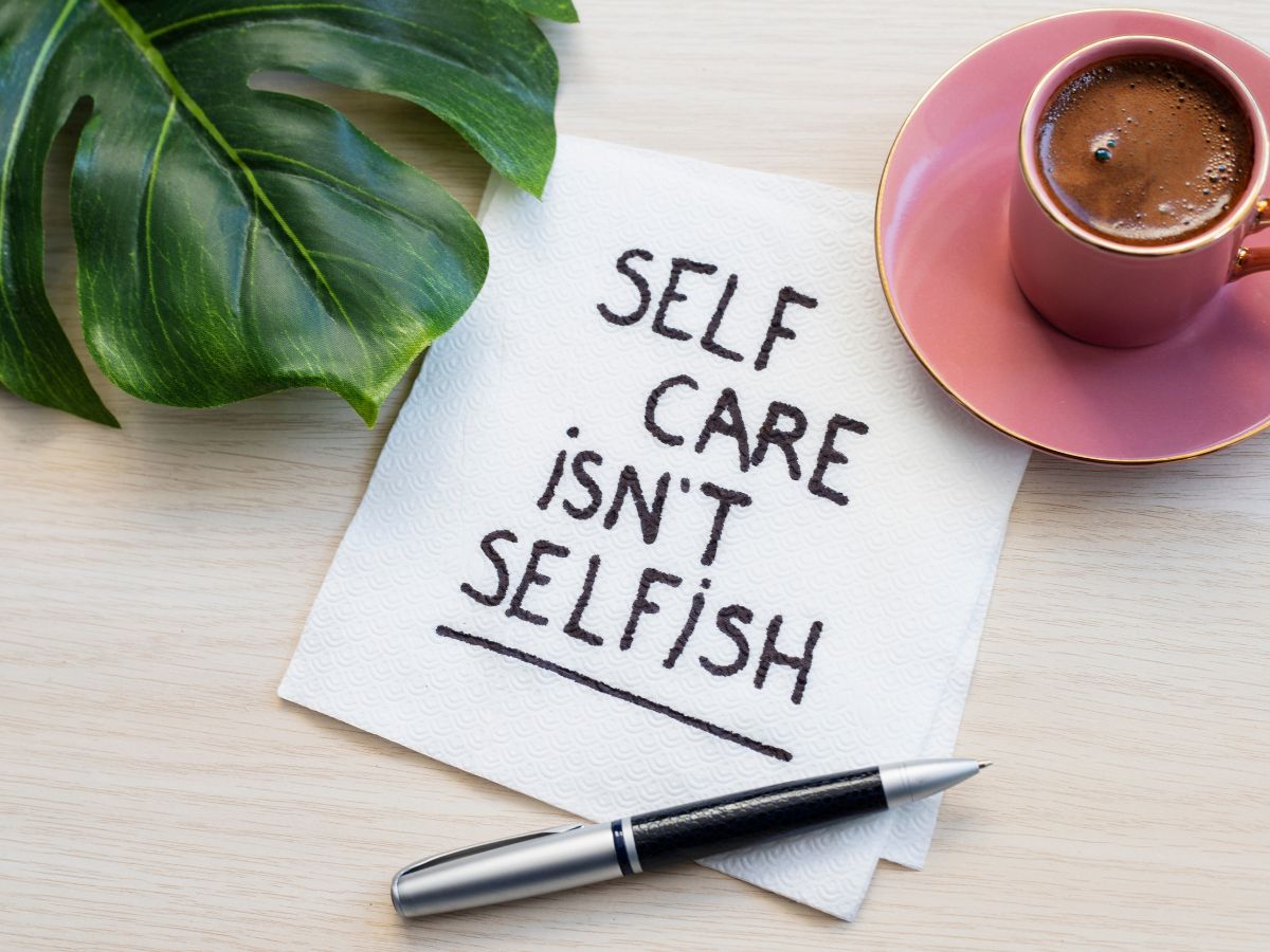 A piece of paper on the table next to a mug and leaf reads "Self care isn't selfish"