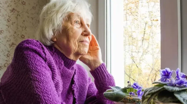 elderly woman staring out window wearing purple jumper