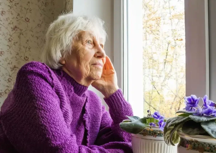 elderly woman staring out window wearing purple jumper