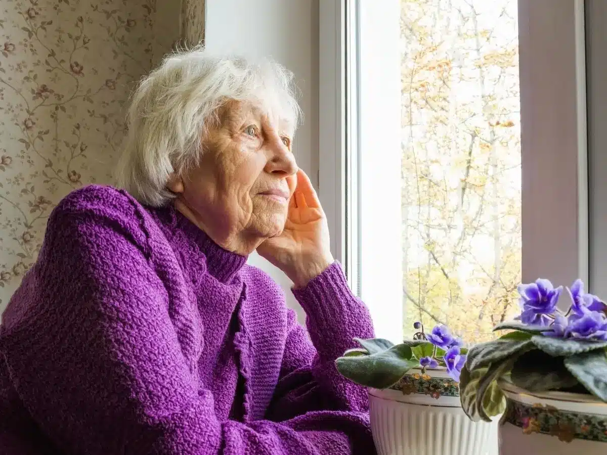 elderly woman staring out window wearing purple jumper