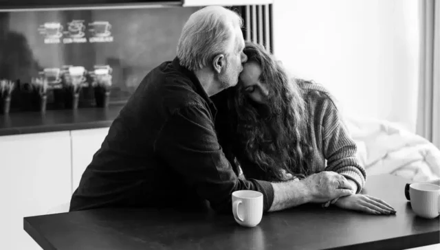 Black and white photo depicting an older man comforting a younger woman, both sat at a kitchen table.