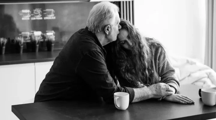 Black and white photo depicting an older man comforting a younger woman, both sat at a kitchen table.