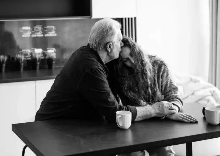 Black and white photo depicting an older man comforting a younger woman, both sat at a kitchen table.