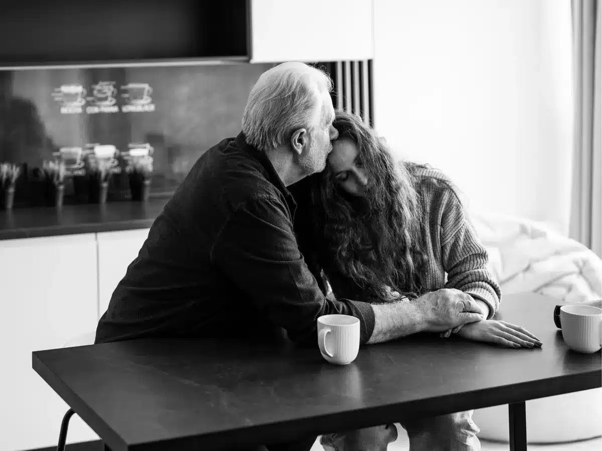 Black and white photo depicting an older man comforting a younger woman, both sat at a kitchen table.