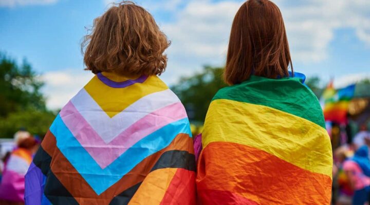 2 people facing away from the camera, noth with different pride flags on their backs. One is the classic rainbow pride flag, the other is the update progressive flag featuring the trans and intersex peoples.