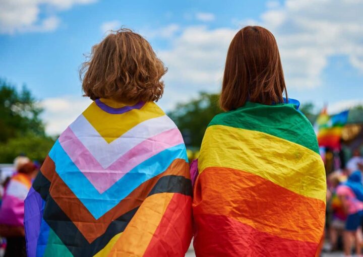 2 people facing away from the camera, noth with different pride flags on their backs. One is the classic rainbow pride flag, the other is the update progressive flag featuring the trans and intersex peoples.