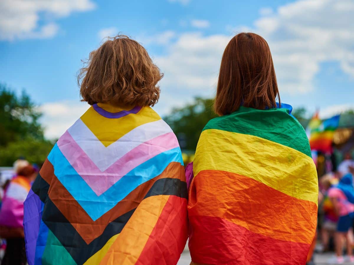 2 people facing away from the camera, noth with different pride flags on their backs. One is the classic rainbow pride flag, the other is the update progressive flag featuring the trans and intersex peoples.